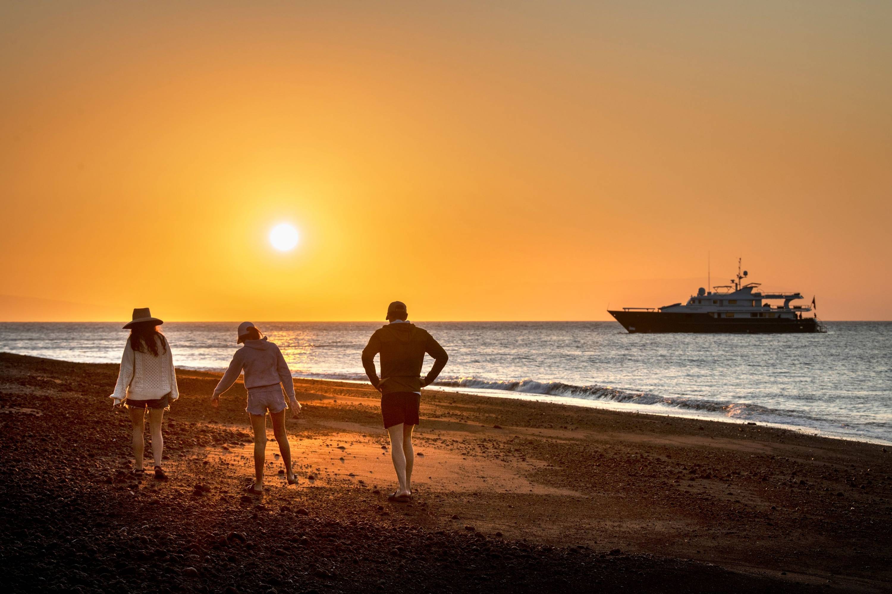 Sonnenuntergang am Pazifikstrand Drei Menschen spazieren bei Sonnenuntergang am Strand der Galapagosinseln, im Hintergrund liegt eine Yacht vor Anker.