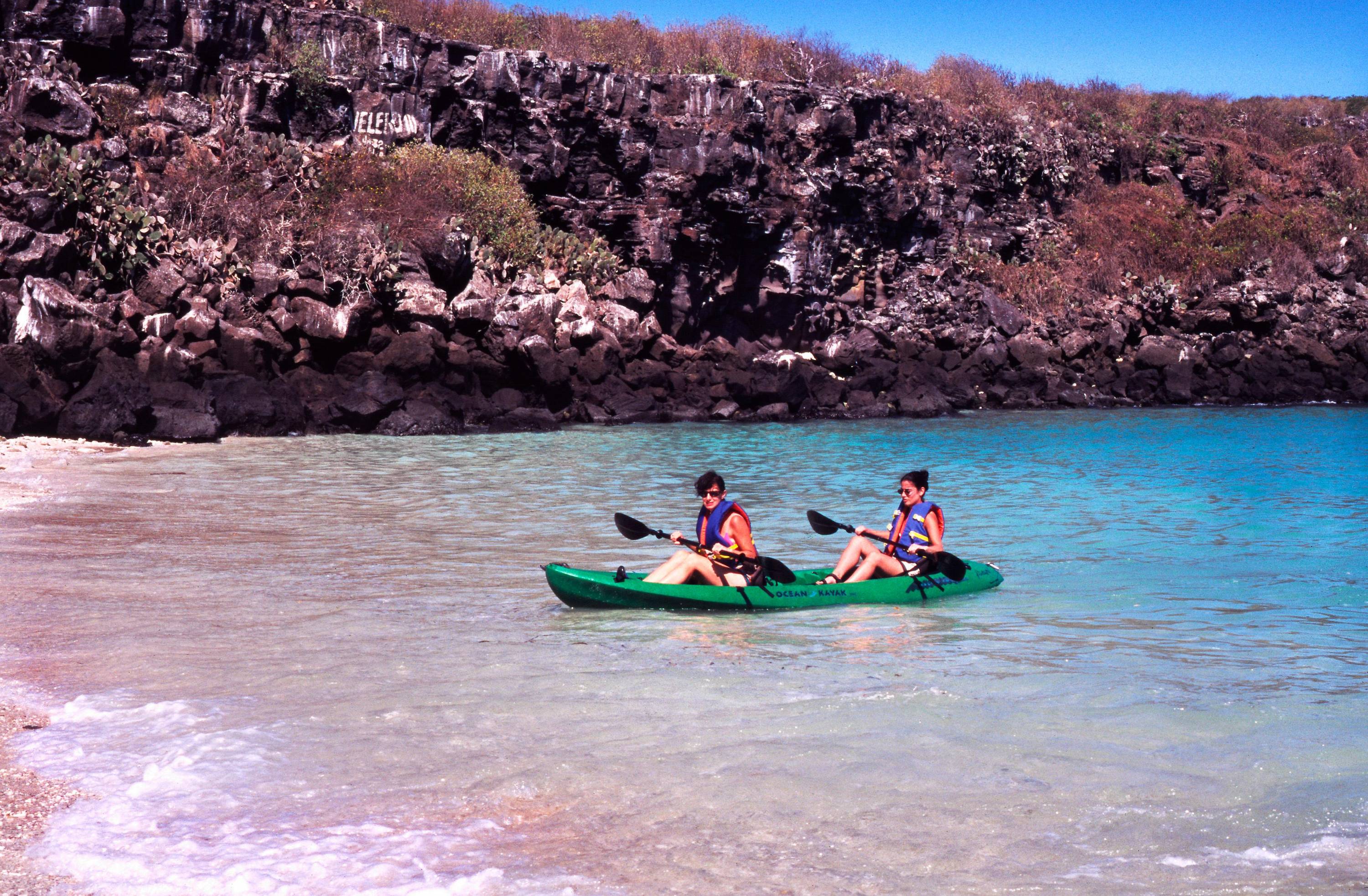 Mit dem Kajak unterwegs im Inselparadies Zwei Personen paddeln in einem grünen Kajak entlang der felsigen Küste einer Galapagos-Insel in türkisblauem Wasser.