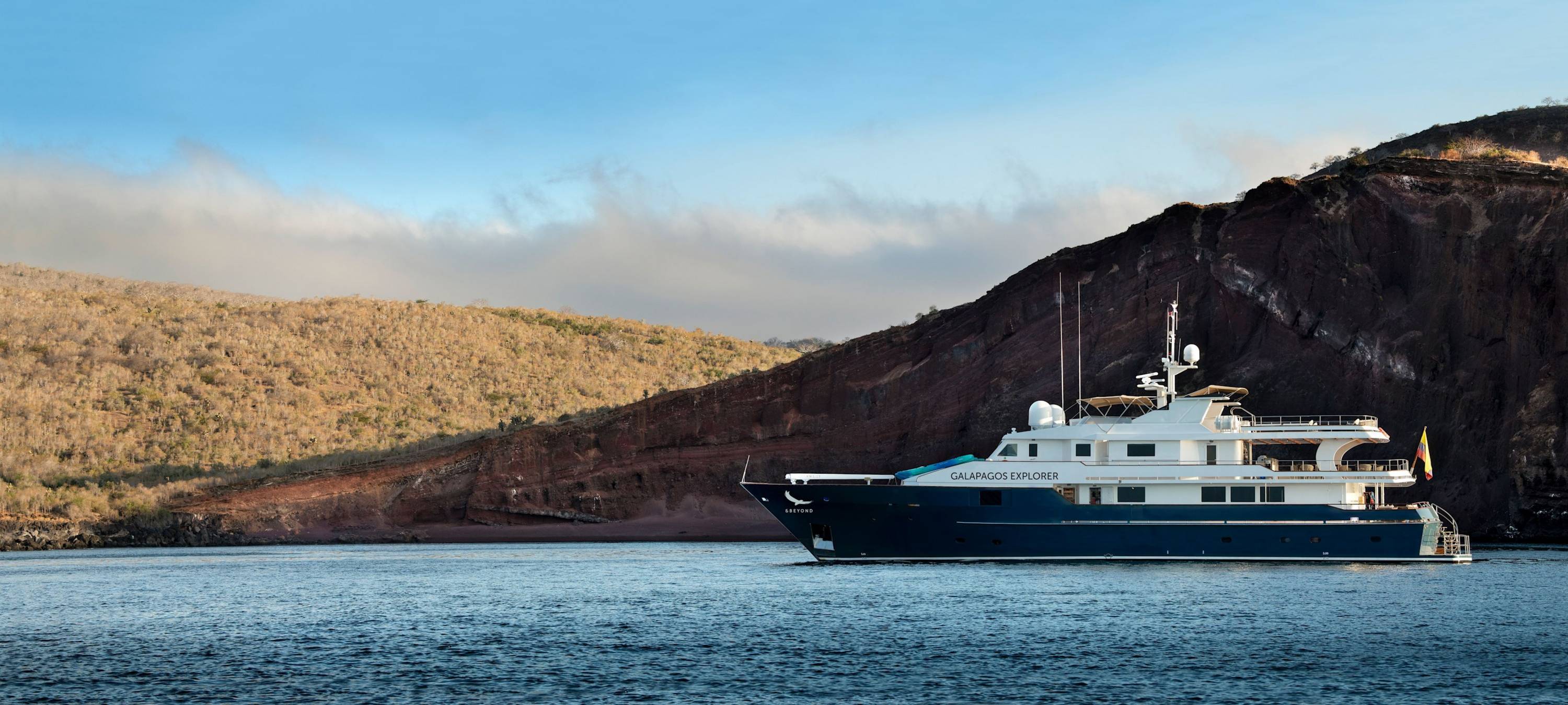 Exklusives Abenteuer auf See Motoryacht „Galapagos Explorer“ liegt vor Anker vor rotbrauner Felsküste auf den Galapagosinseln bei ruhiger See im Morgen- oder Abendlicht.