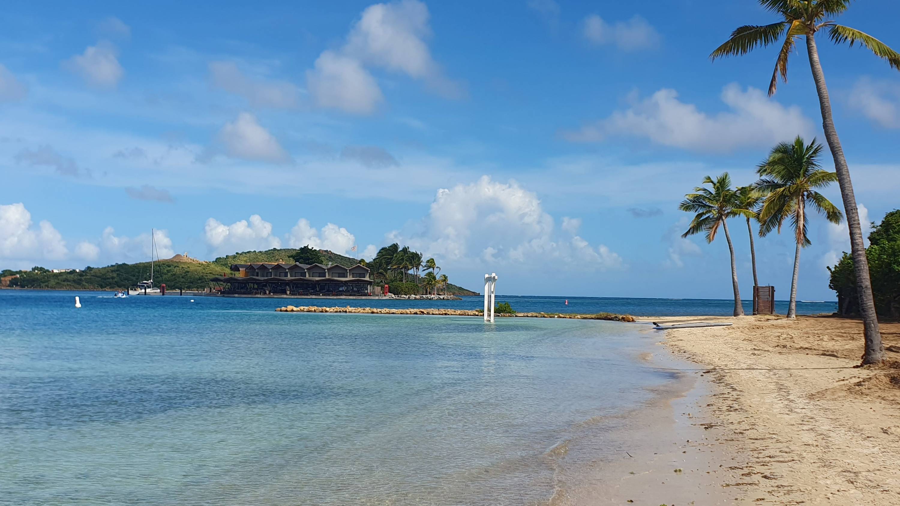 Tropischer Sandstrand mit Palmen und flach abfallendem Wasser, im Hintergrund luxuriöse Stelzenbungalows und ein Segelboot in einer ruhigen Lagune