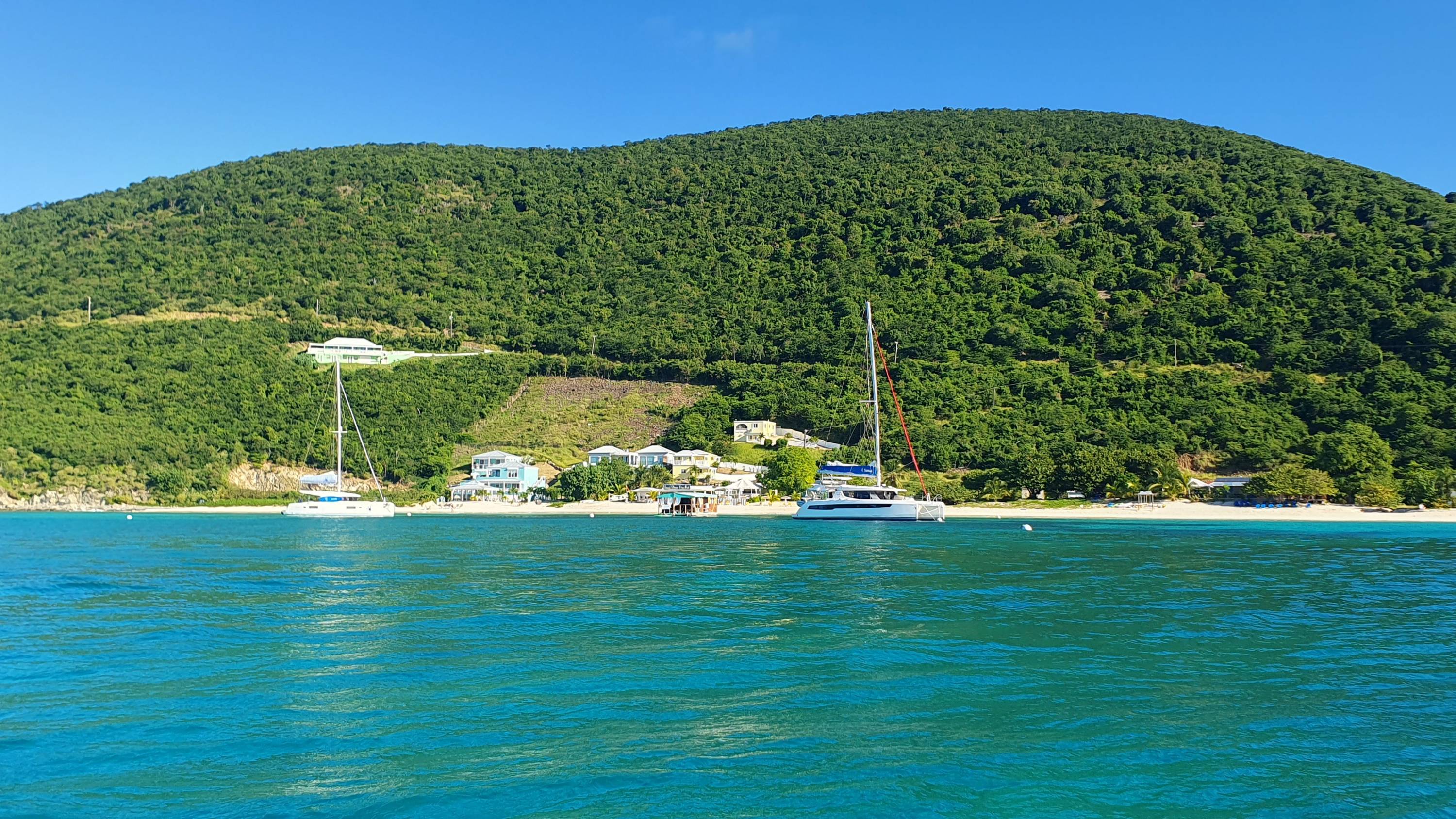 Zwei Katamarane ankern vor einem tropischen Strand auf der Britischen Jungferninseln mit grüner Hügellandschaft im Hintergrund.