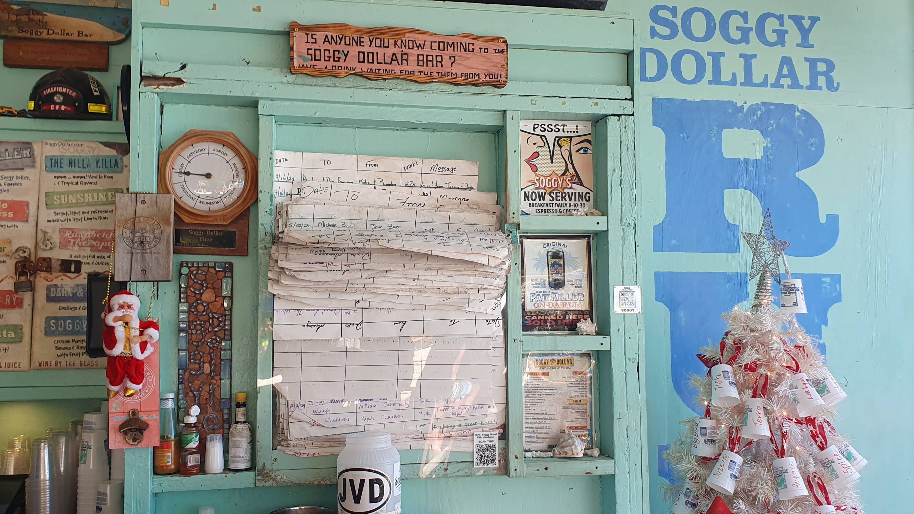 Innenaufnahme der Soggy Dollar Bar mit türkisfarbener Wand, improvisierter Nachrichtentafel, Santa-Figur und weißem Plastikbecher-Weihnachtsbaum.