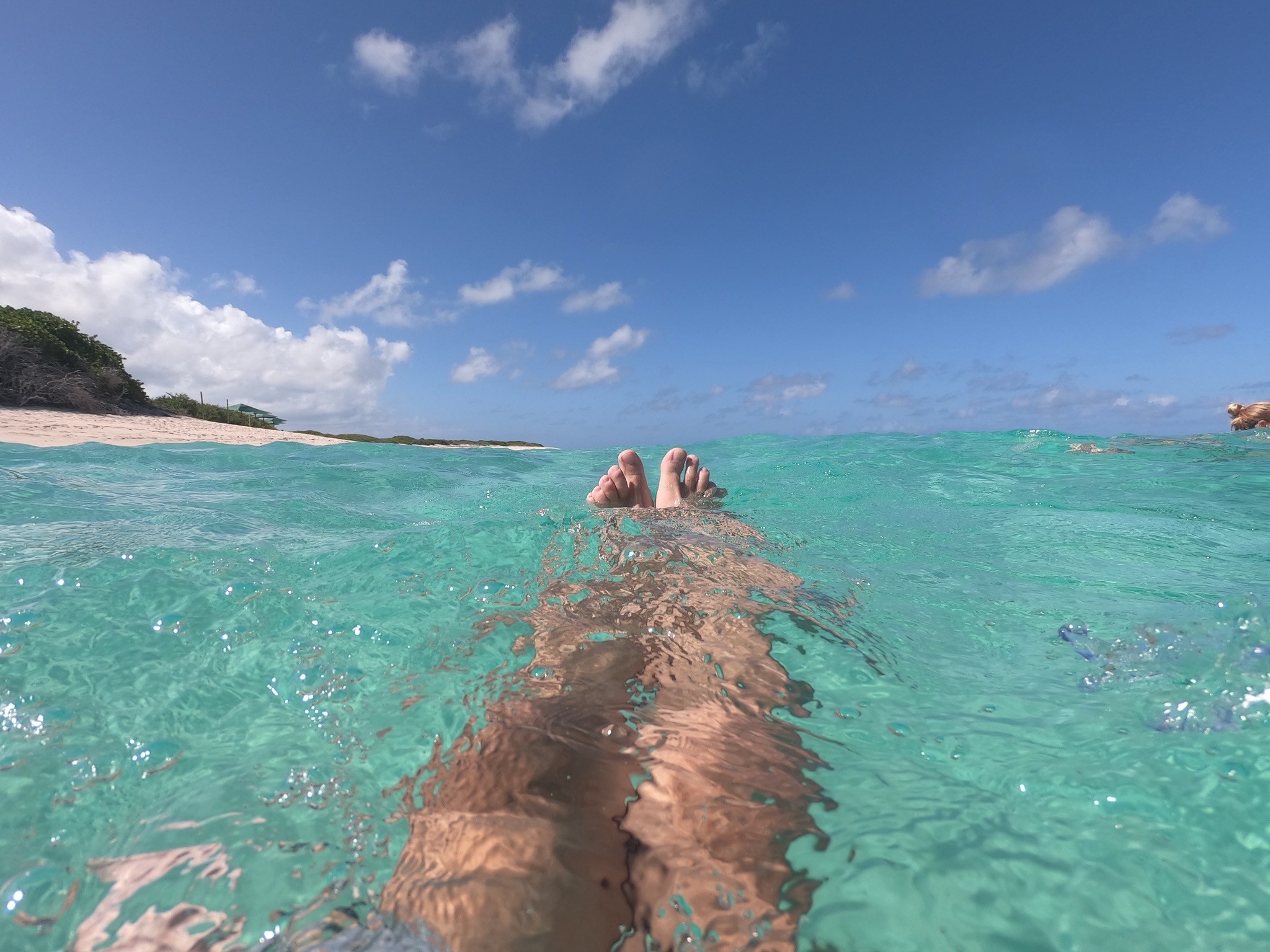 Perspektive aus dem Wasser auf ausgestreckte Beine in türkisblauer Lagune vor tropischem Sandstrand unter blauem Himmel