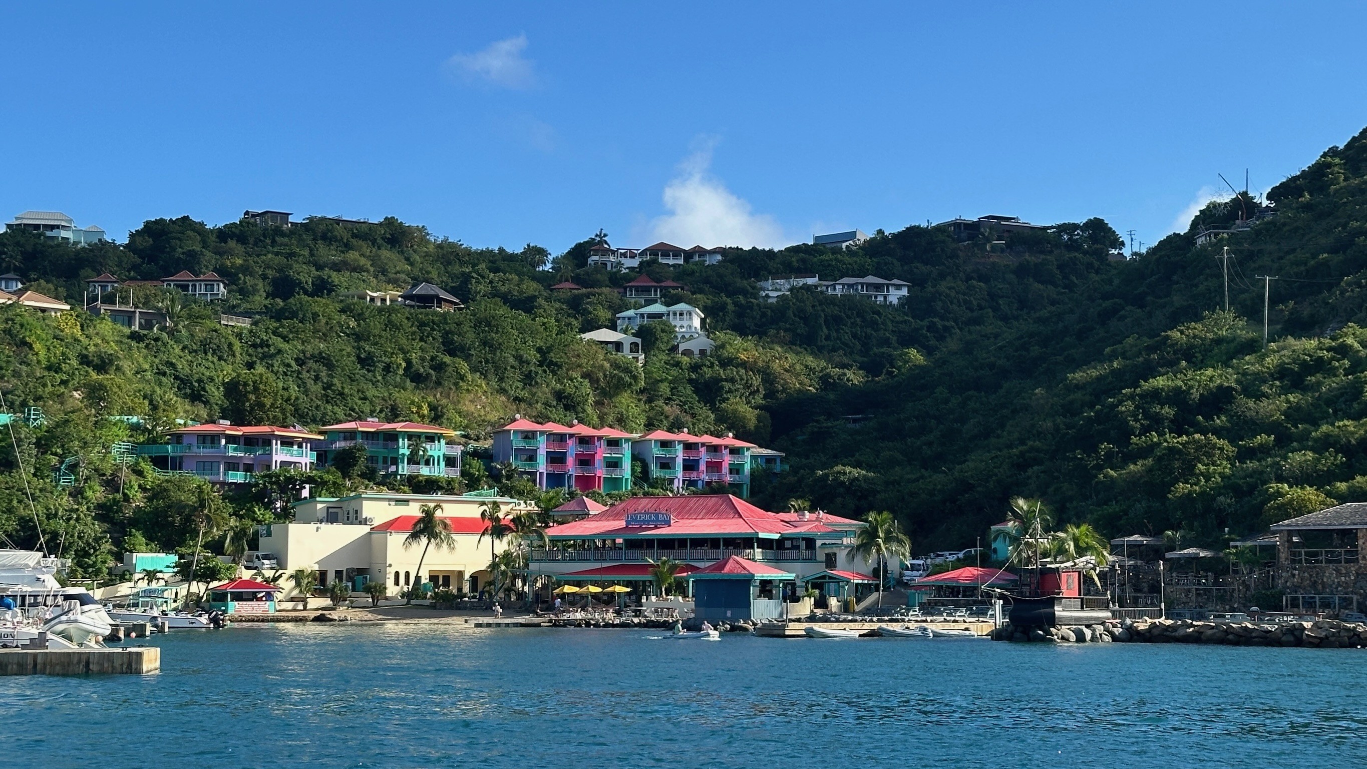 Bunter Hotelkomplex und Uferpromenade mit karibischen Gebäuden an einem grünen Hang, aufgenommen vom Wasser aus