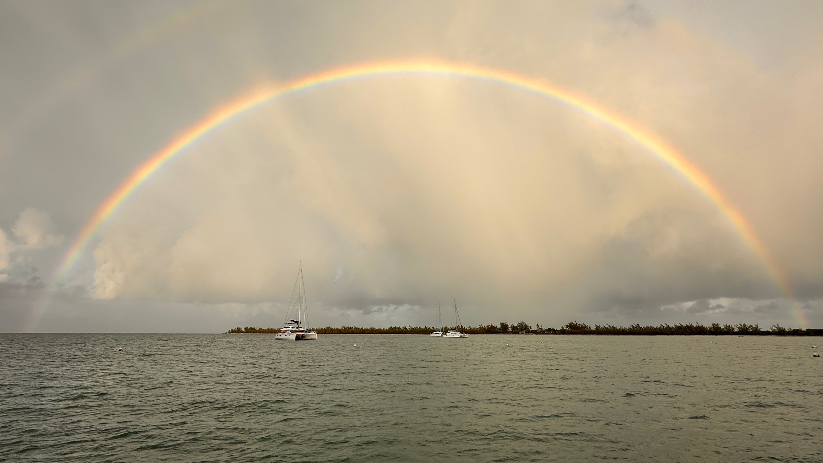 Vollständiger Regenbogen spannt sich über ein ruhiges Meer mit ankernden Segelbooten und einer flachen Insel am Horizont