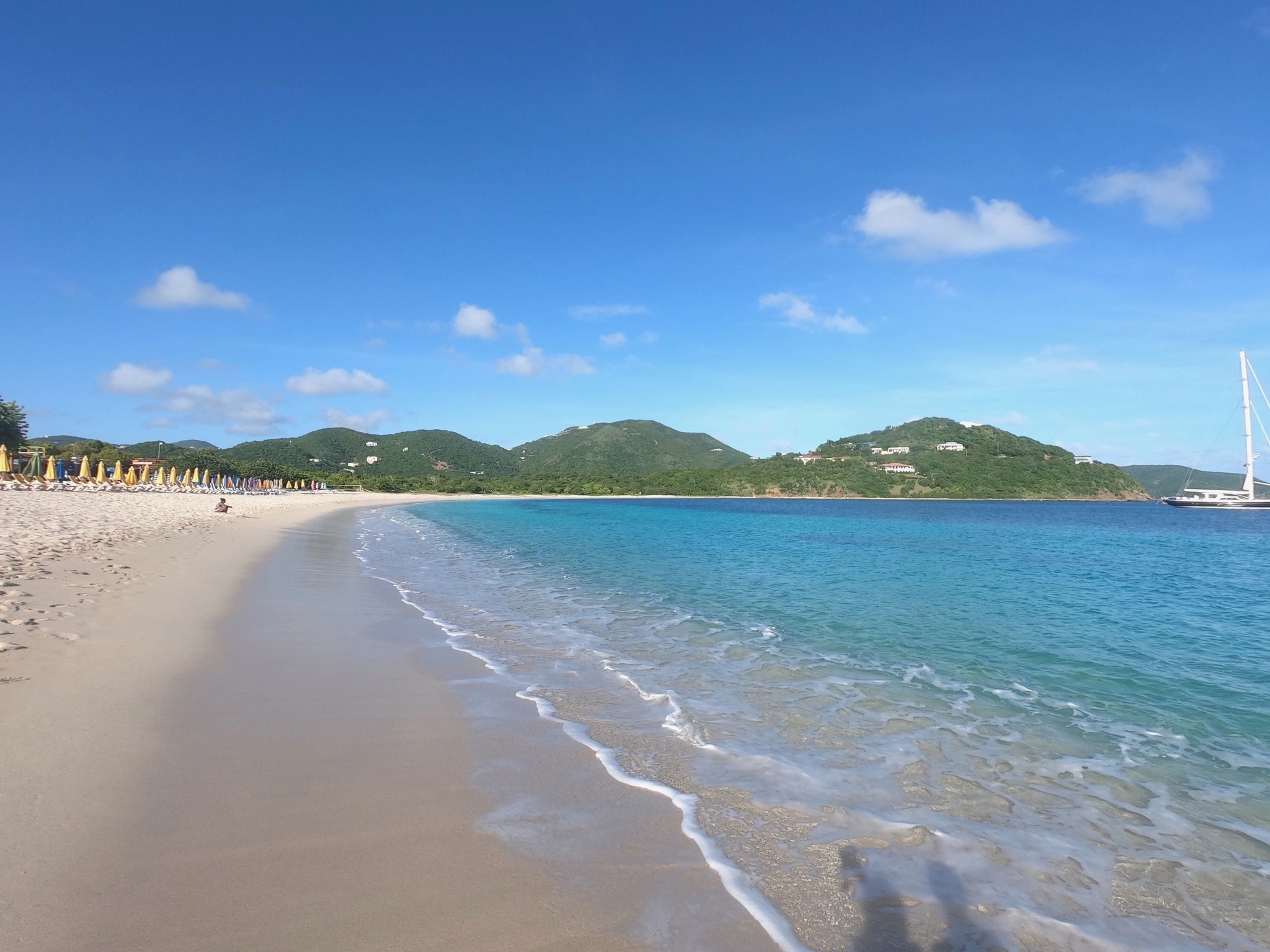 Weiter Sandstrand mit türkisblauem Meer, Sonnenschirmen und einer Yacht vor grüner Inselkulisse