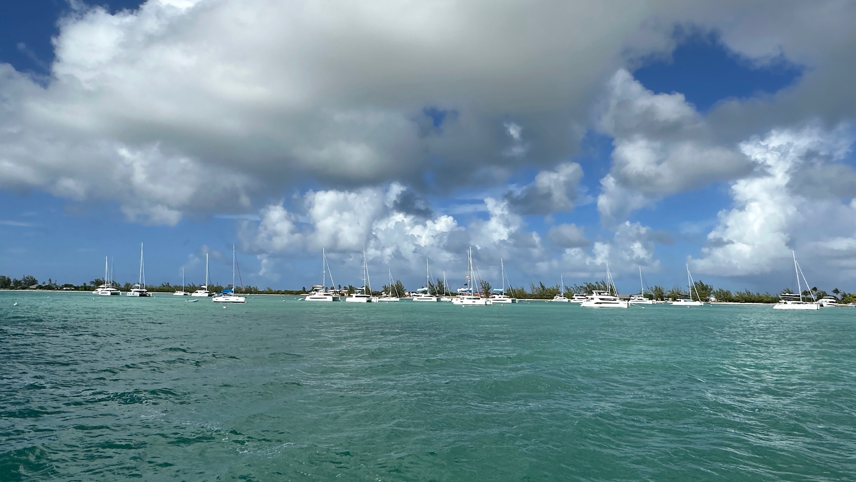 Zahlreiche Yachten und Katamarane ankern vor einer flachen Insel unter eindrucksvollen Wolkenformationen und blauem Himmel