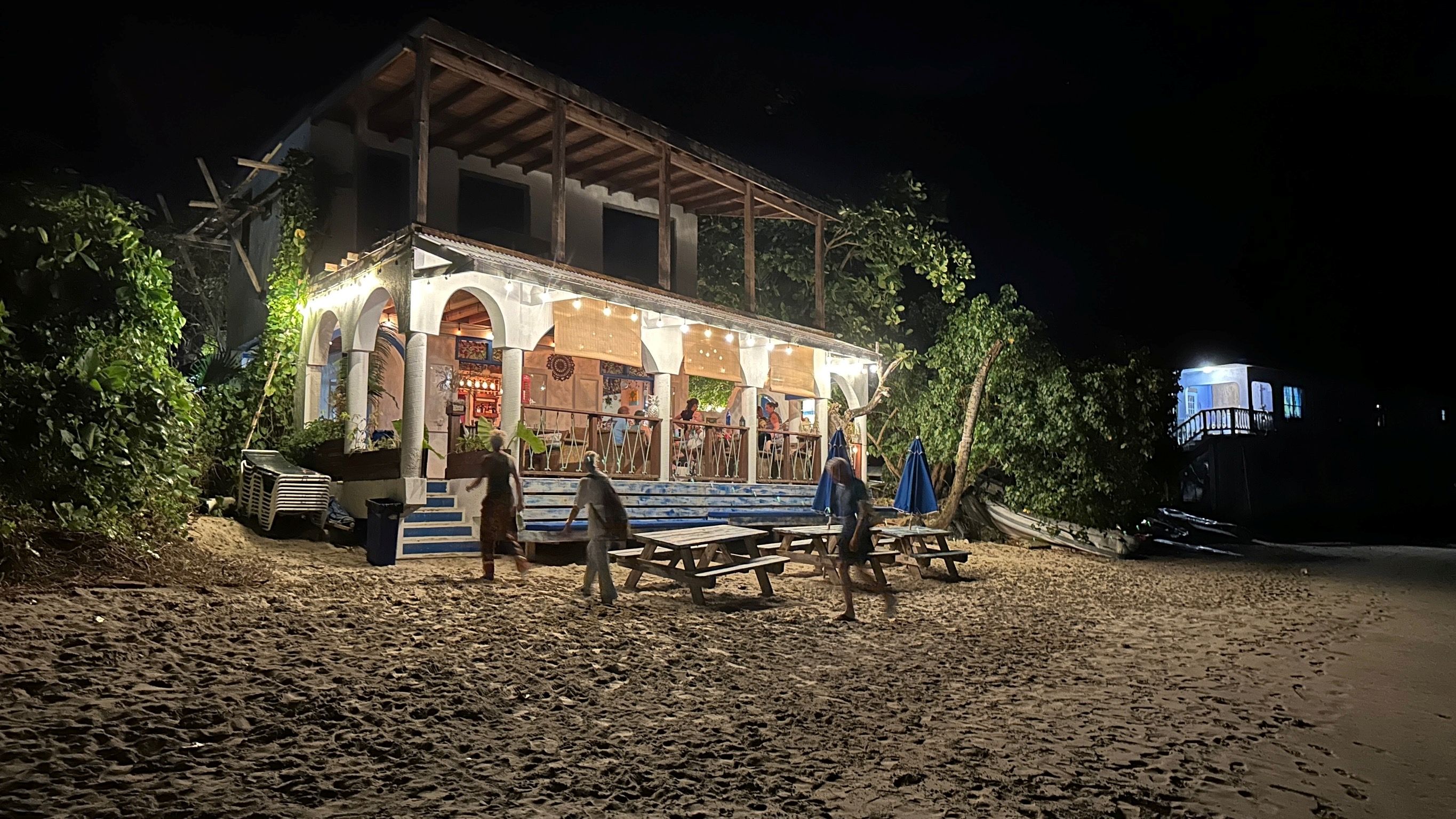 Kleines, beleuchtetes Strandrestaurant bei Nacht mit Menschen und Palmen im Hintergrund