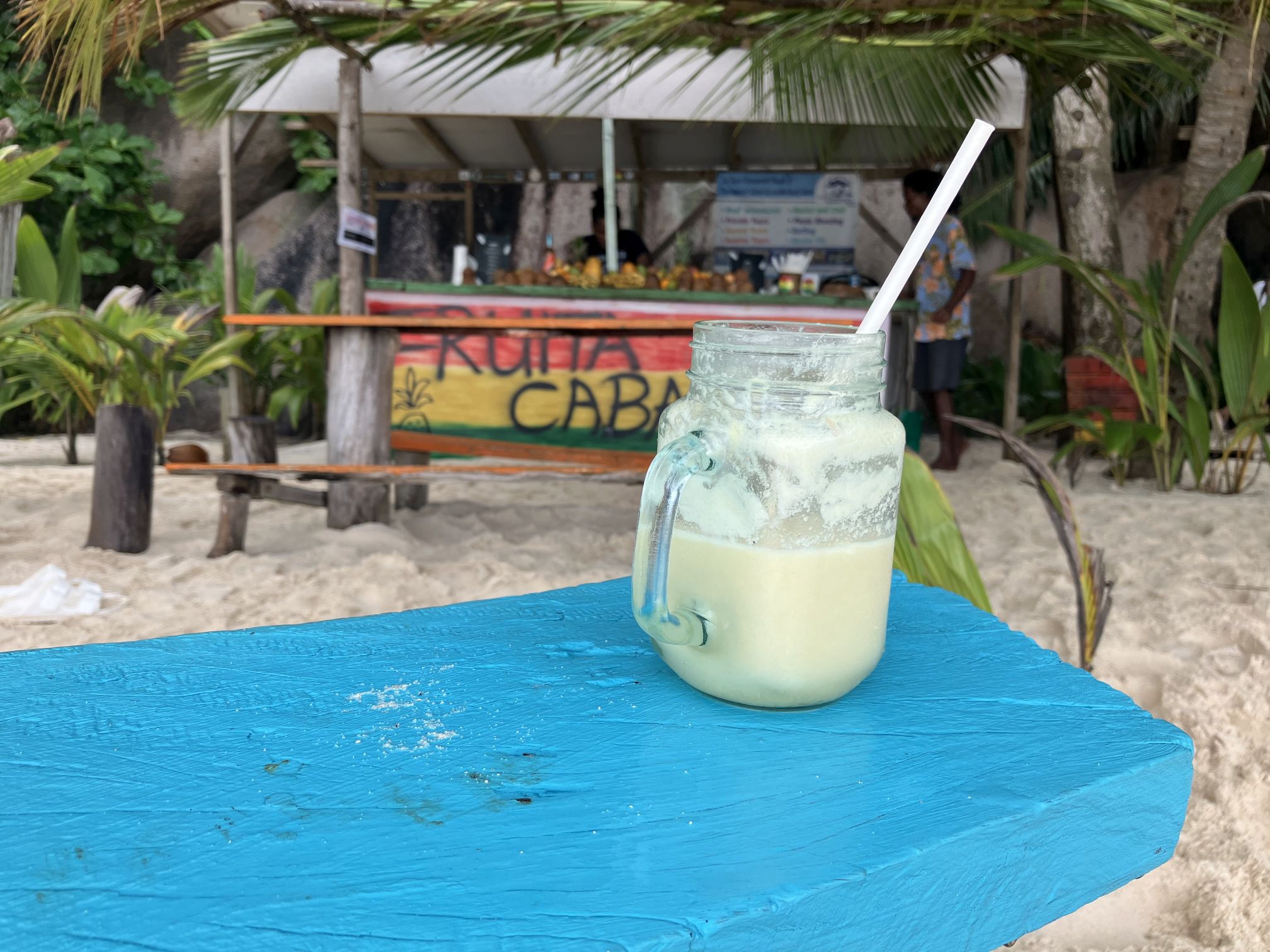 Cocktailglas mit Strohhalm steht auf blau gestrichenem Holztisch vor einer Strandbar.
