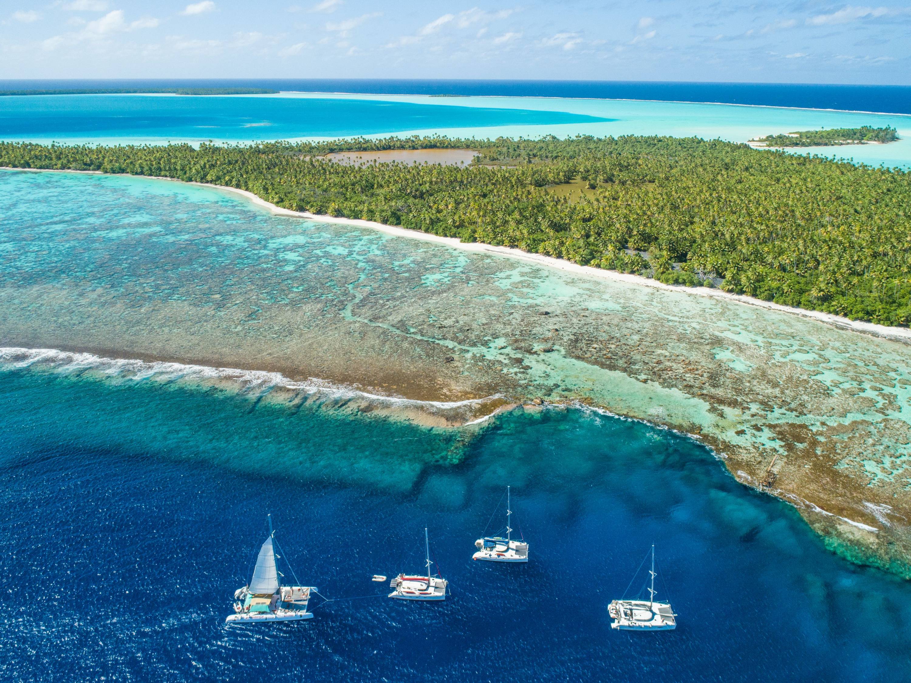 Luftaufnahme von Segelkatamaranen vor dem Riffgürtel der Insel Tetiaroa mit üppiger Vegetation und türkisfarbener Lagune im Hintergrund.