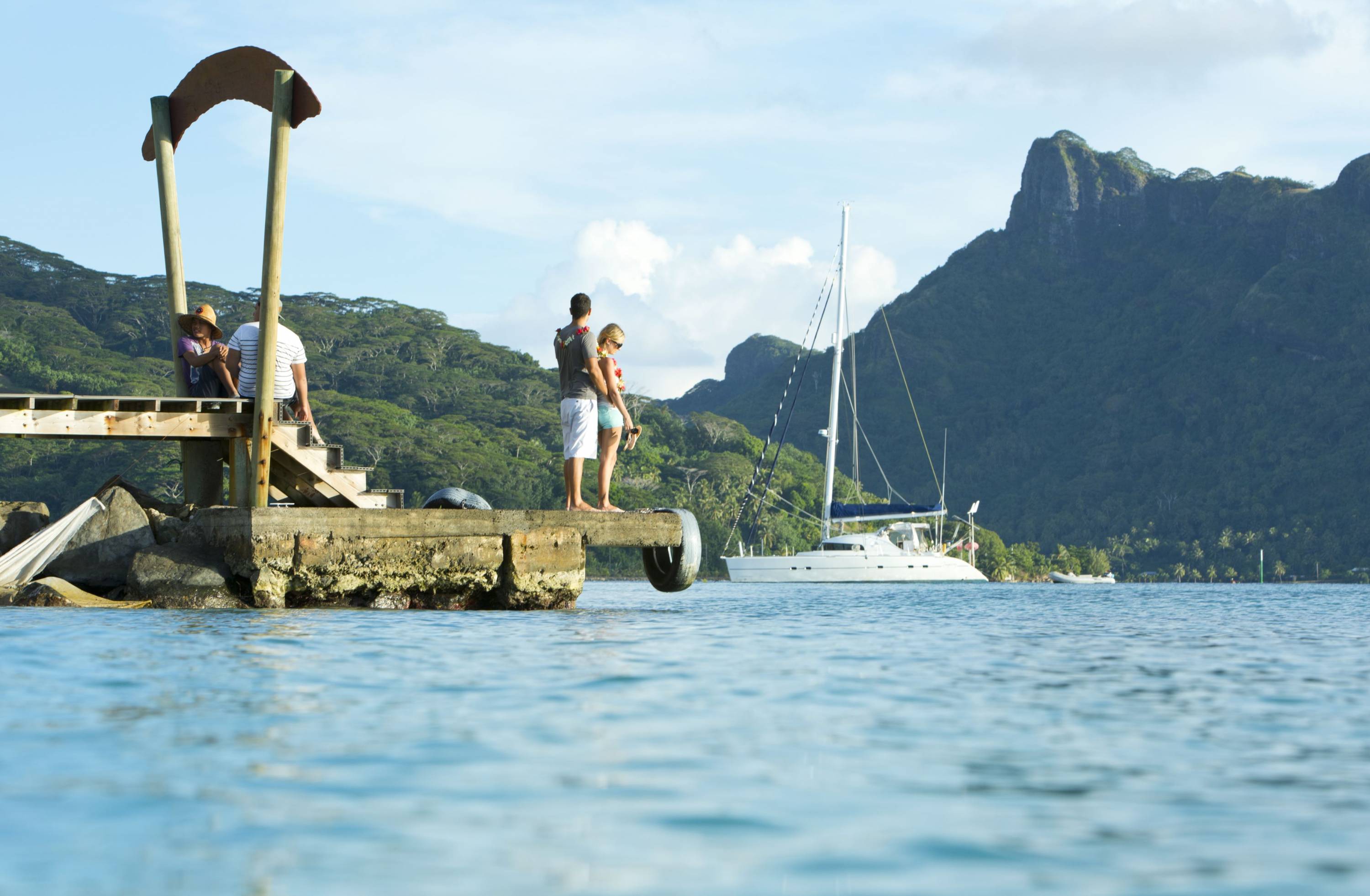 Menschen stehen auf einem Steg auf Huahine mit Blick auf ein Segelboot vor der grünen Berglandschaft der Insel.