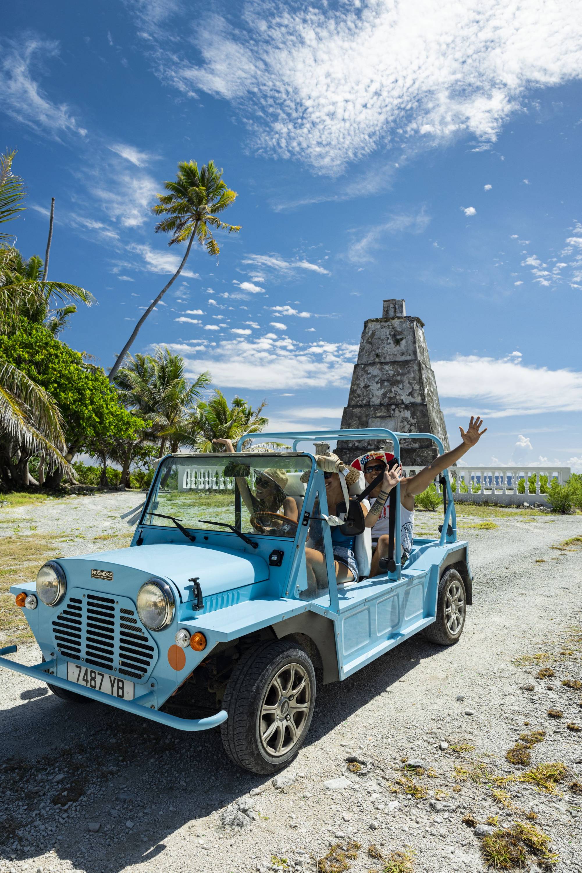 Fröhliche Reisende in einem blauen Mini-Moke auf einer Erkundungstour durch Fakarava, vorbei an einem historischen Denkmal.